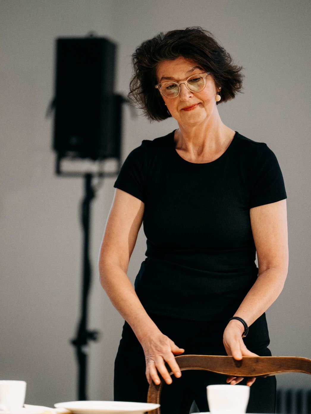 Eine Frau richtet ruhig und konzentriert einen Stuhl an einem gedeckten Tisch aus. Ihr Blick ist nach unten gerichtet, die Bewegung wirkt bedacht und sorgfältig. Die schlichte Umgebung und das reduzierte Setting lenken den Fokus auf die kleine, alltägliche Handlung, die eine stille, beinahe inszenierte Atmosphäre erzeugt.