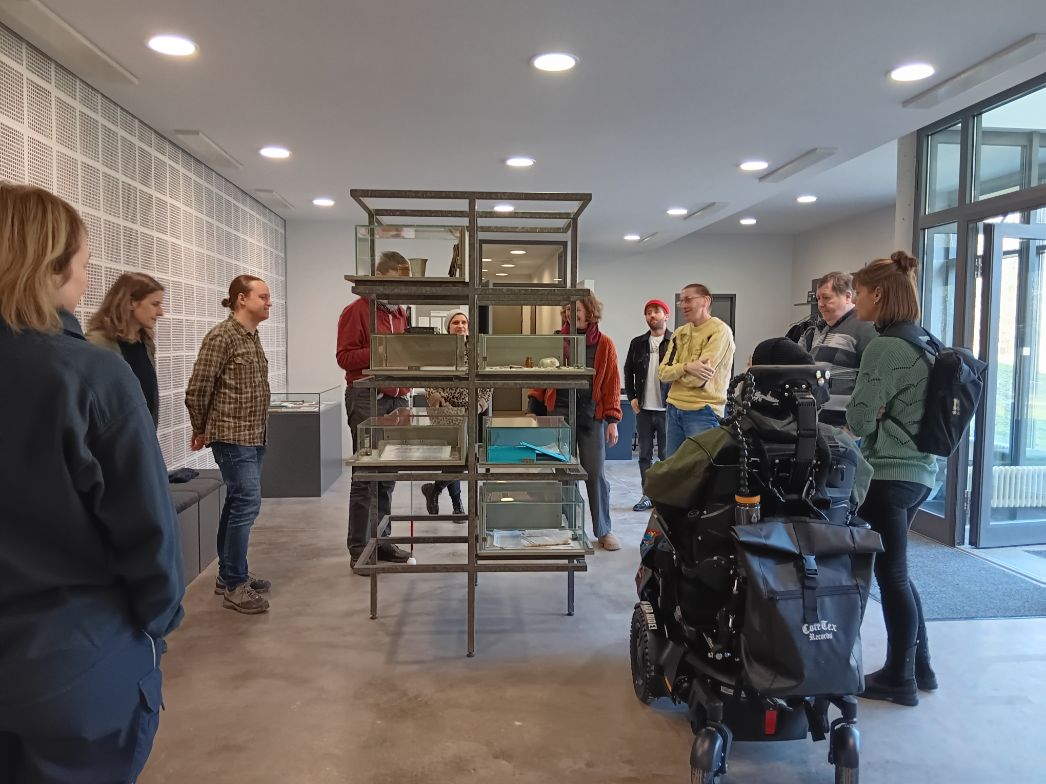 Gruppe von Menschen in einem modernen Ausstellungsraum, die um eine zentrale Vitrine stehen und sich austauschen. In der Mitte befindet sich ein mehrstöckiges Metallregal mit Glasvitrinen, in denen Objekte ausgestellt sind. Die Teilnehmenden wirken interessiert und im Gespräch, einige lächeln. Eine Person sitzt in einem elektrischen Rollstuhl. Der Raum ist hell, mit großen Fenstern, schlichten Wänden und gleichmäßig verteilten Deckenleuchten.