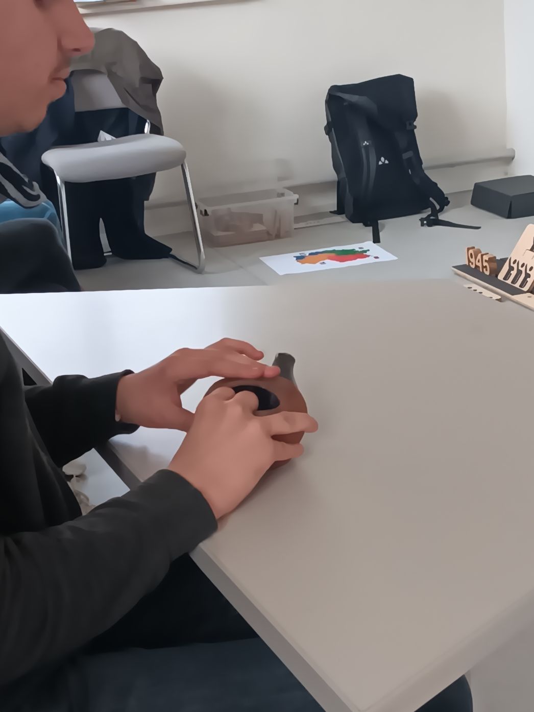 Eine Person sitzt an einem Tisch und hält mit beiden Händen ein kleines, rundliches Objekt aus dunklem Material. Im Hintergrund sind ein Rucksack, ein Stuhl und eine auf dem Boden liegende Karte zu sehen. Rechts am Tischrand befindet sich ein Holzobjekt mit der Jahreszahl „1945“.
