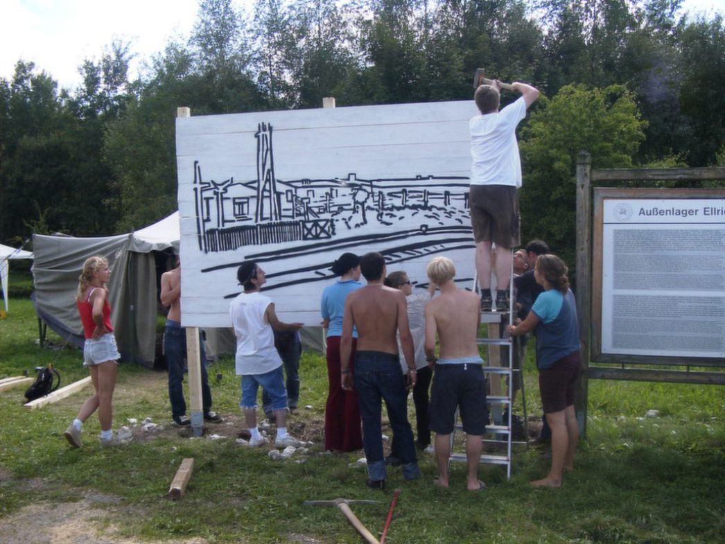Eine Gruppe junger Menschen arbeitet gemeinsam an einer großen Tafel im Freien, auf die eine historische Szene gezeichnet wird. Die Aufnahme zeigt ein Workcamp bzw. gemeinschaftliches Erinnerungsprojekt an einem Ort eines ehemaligen Außenlagers.