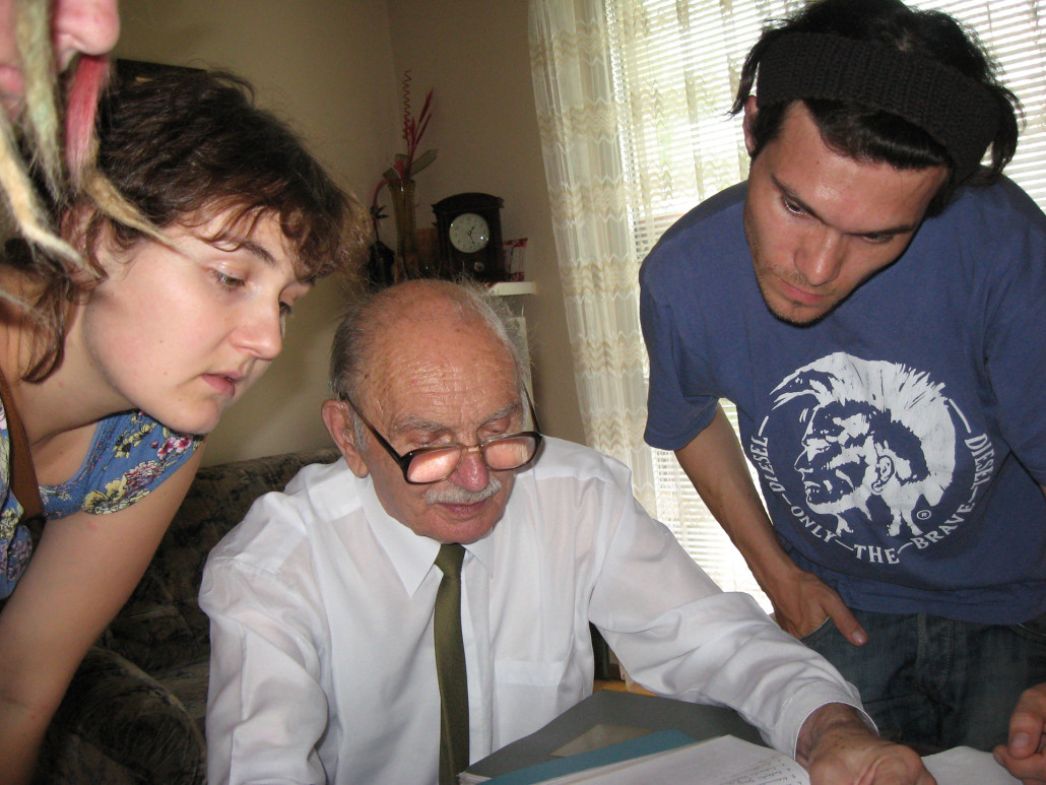 Ein älterer Mann sitzt an einem Tisch und zeigt zwei jüngeren Personen Dokumente. Die Szene vermittelt ein Gespräch bzw. Zeitzeugenaustausch, bei dem Erfahrungen und Erinnerungen weitergegeben werden.