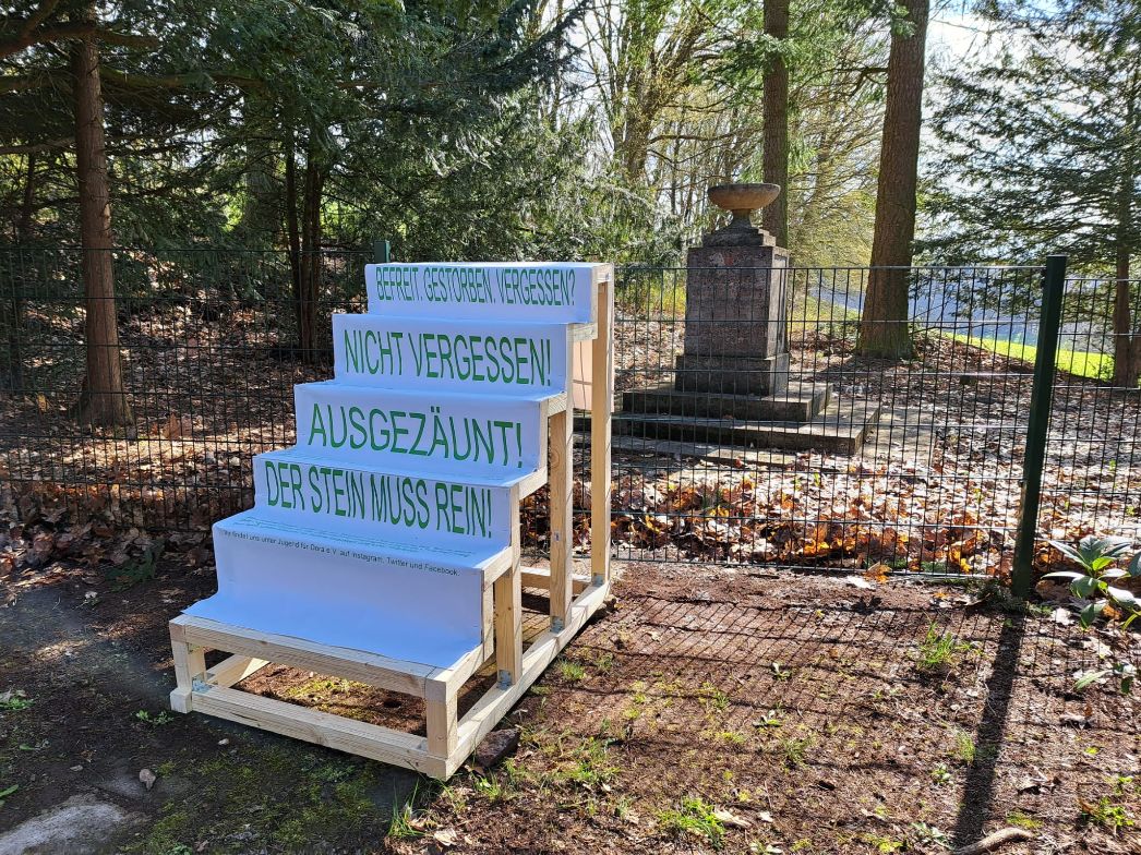 Eine temporäre Installation vor einem umzäunten Denkmal. Auf gestapelten Tafeln stehen Forderungen wie „Nicht vergessen!“ und „Der Stein muss rein!“, die auf Erinnerung und den Umgang mit dem Gedenkort aufmerksam machen. Die Tafeln bilden eine Treppe, welche die Höhe des Zaunes erreicht.