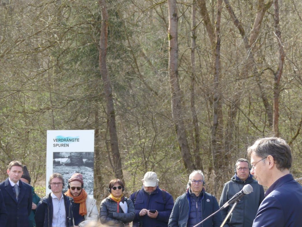 Gruppe von Menschen im Freien, die einer Rede zuhören. Rechts steht ein Mann im Anzug an einem Mikrofon und spricht. Die Zuhörenden stehen aufmerksam davor, einige mit Kopfhörern oder Blick auf ihre Notizen. Hinter der Gruppe befindet sich eine Informationstafel mit der Überschrift „Verdrängte Spuren“. Die Umgebung ist von Bäumen geprägt und wirkt wie ein Gedenk- oder Ausstellungsort im Wald.
