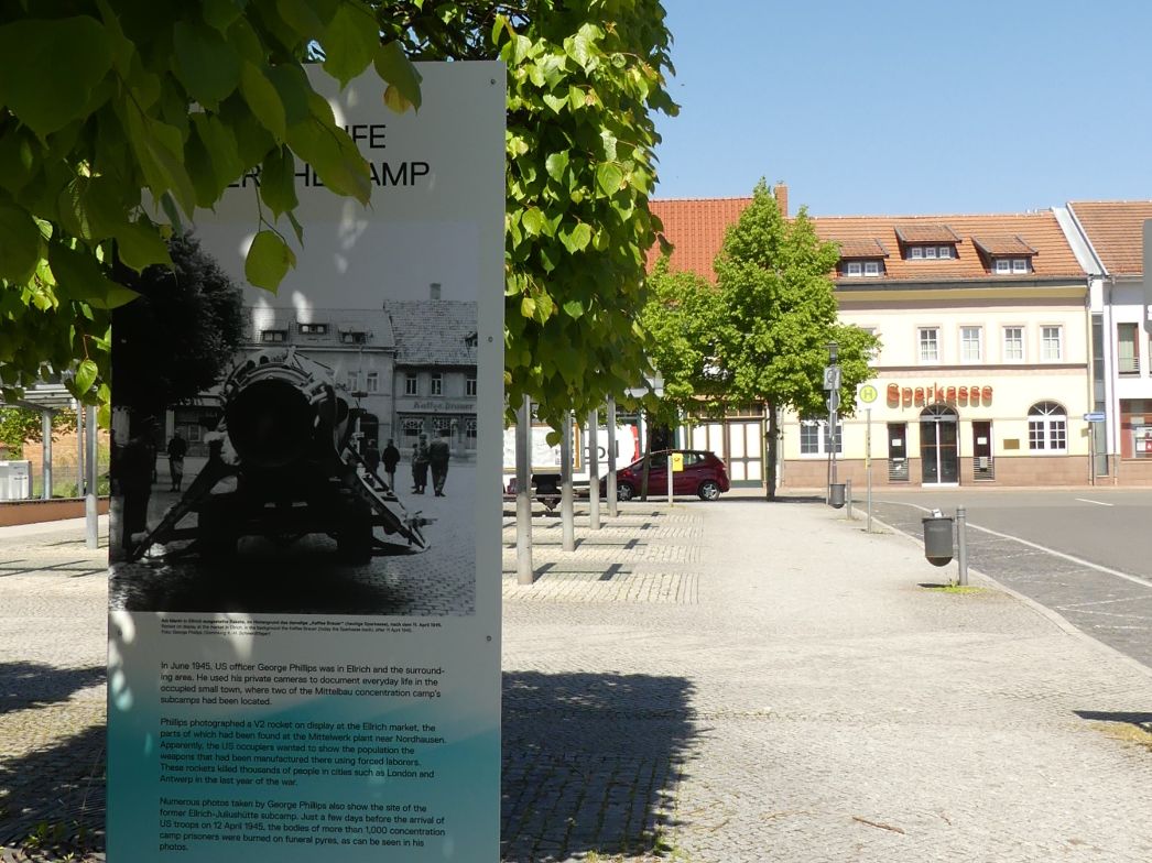 Freistehende Informationstafel im öffentlichen Raum, teilweise von grünen Blättern beschattet. Die Tafel zeigt ein historisches Schwarzweißfoto und erläuternden Text auf Englisch zum Thema Mittelbau-Dora und dem Außenlager Ellrich. Im Hintergrund ist ein ruhiger Marktplatz mit gepflastertem Boden, Bäumen und Gebäuden zu sehen, darunter ein Haus mit der Aufschrift „Sparkasse“.