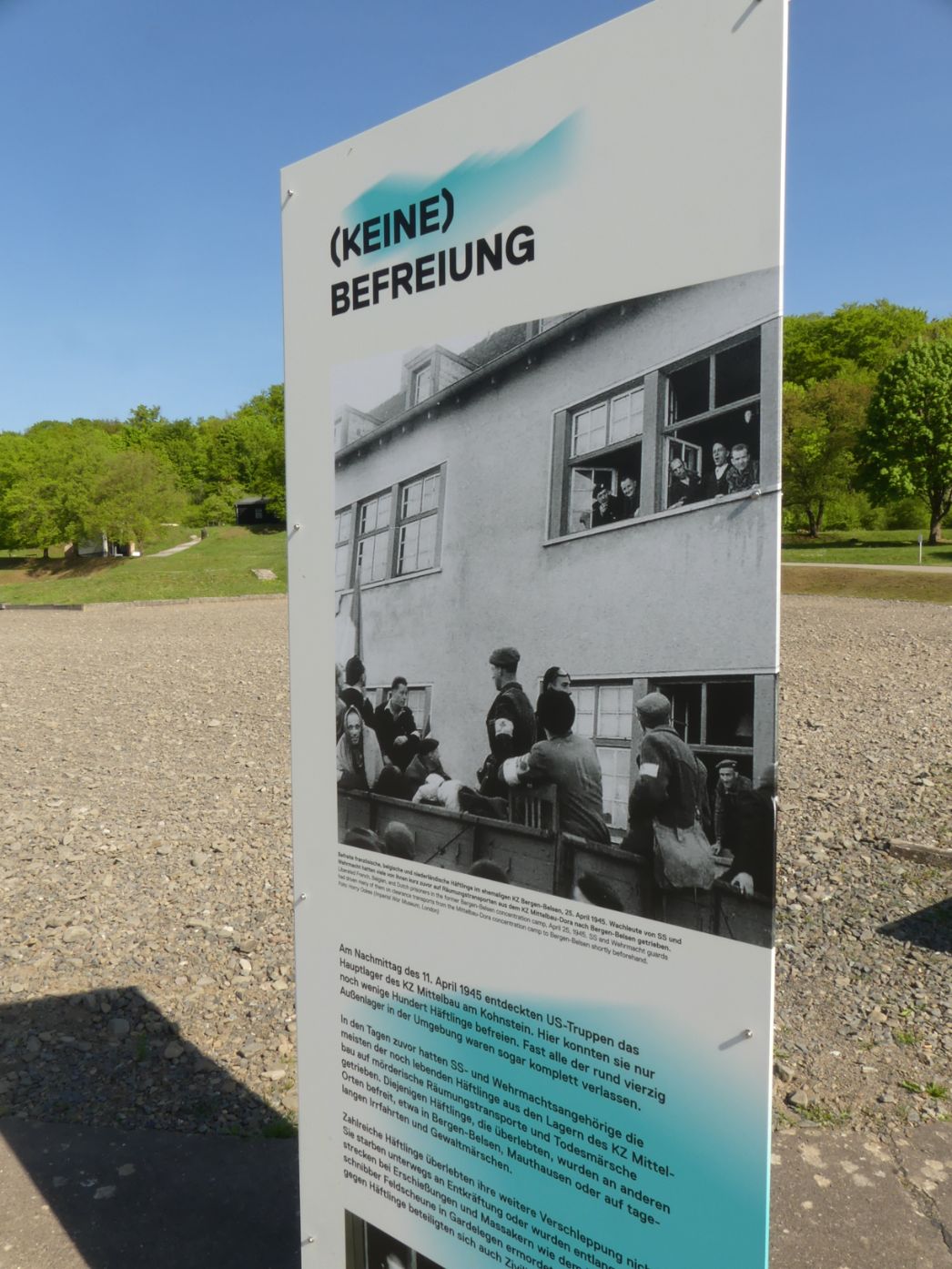 Eine Ausstellungstafel im Außenbereich mit dem Titel „(KEINE) BEFREIUNG“. Sie zeigt ein historisches Foto und erklärt die Situation im April 1945: Viele Häftlinge des KZ Mittelbau wurden nicht sofort befreit, sondern zuvor verlegt oder auf Todesmärsche geschickt.