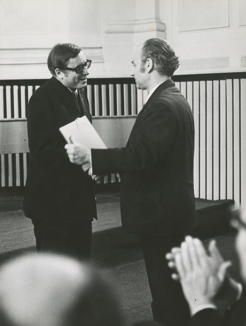Schwarzweißfotografie eines Mannes mittleren Alters mit Brille, der an einem Schreibtisch sitzt und in ein aufgeschlagenes Buch schreibt oder liest. Er trägt ein Jackett und wirkt konzentriert bei der Arbeit. Auf dem Tisch liegen weitere Bücher und Papiere. Im Hintergrund sind Regale oder ein Arbeitsraum zu erkennen, was auf eine Schreib- oder Büroumgebung hinweist.