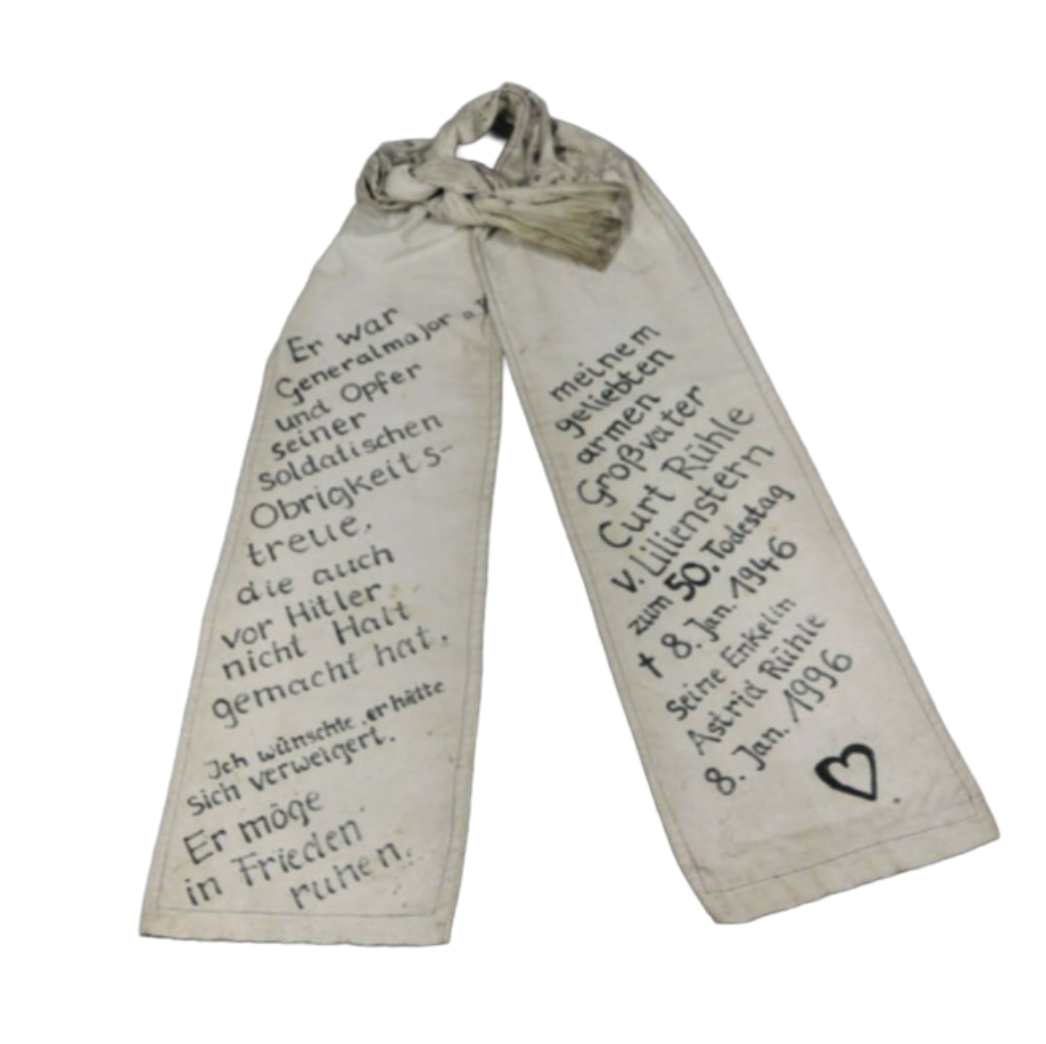 Zwei lange, schmale Stoffbänder, oben zu einer Schleife zusammengebunden. Auf beiden Bändern steht handschriftlicher Text in deutscher Sprache. Die Inschrift erinnert an eine verstorbene Person und enthält persönliche Worte sowie Daten. Am unteren Ende eines Bandes ist ein kleines gezeichnetes Herz zu sehen.