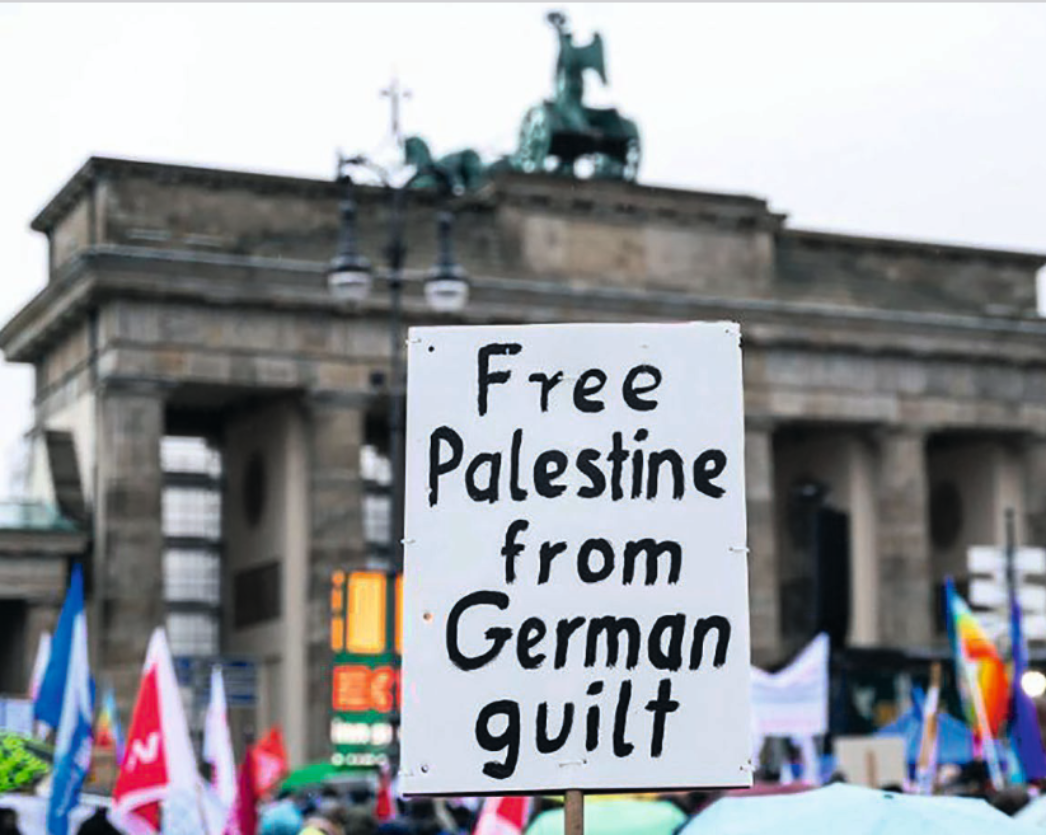 Ein Demonstrationsschild vor dem Brandenburger Tor mit der Aufschrift „Free Palestine from German guilt“, gehalten in einer Menschenmenge mit Fahnen im Hintergrund.