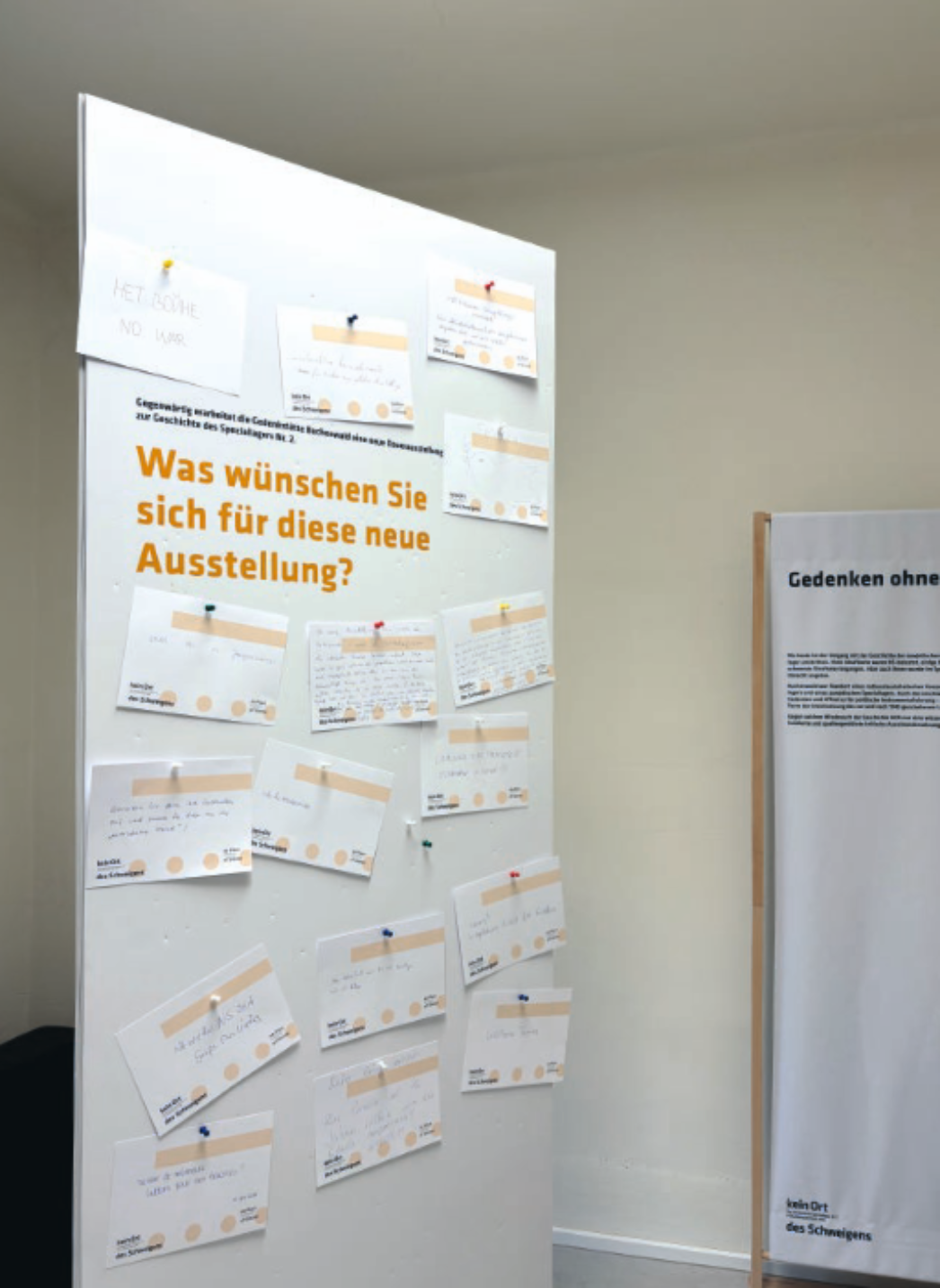 Weiße Pinnwand mit der Überschrift „Was wünschen Sie sich für diese neue Ausstellung?“. Darauf sind zahlreiche handschriftlich ausgefüllte Karten mit Kommentaren und Wünschen von Besucherinnen und Besuchern befestigt. Rechts daneben steht eine weitere Informationstafel mit Text zur Ausstellung. Der Raum ist schlicht und hell gestaltet.