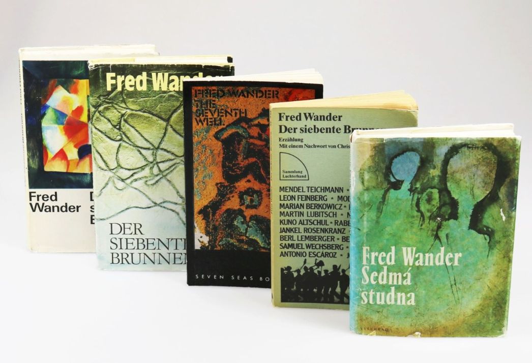 Mehrere Ausgaben eines Buches von Fred Wander sind nebeneinander aufgestellt. Die unterschiedlich gestalteten Buchcover zeigen verschiedene Farben und Illustrationen, darunter abstrakte Motive und Figuren. Auf einigen Covern ist der Titel „Der siebente Brunnen“ in deutscher Sprache zu lesen, auf einem anderen in einer fremdsprachigen Variante. Die Bücher stehen leicht versetzt vor einem neutralen, hellen Hintergrund.