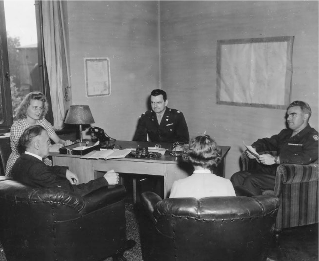 Schwarz-weiß-Fotografie eines Büros: Ein US-amerikanischer Offizier sitzt hinter einem Schreibtisch und spricht mit drei zivilen Personen, die ihm gegenüber in Sesseln sitzen, sowie einem weiteren Soldaten. Auf dem Tisch liegen Dokumente und ein Telefon. Die Szene zeigt eine Besprechung zwischen Militärverwaltung und deutscher Zivilbevölkerung nach Kriegsende.