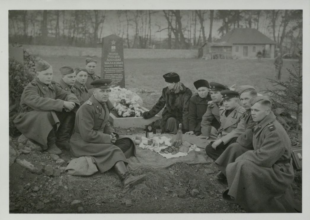 Historische Schwarzweißaufnahme einer Gruppe von Menschen in militärischer Kleidung, die um ein Grab im Freien sitzen. In der Mitte liegt auf einer Decke Essen und Getränke, als würde ein gemeinsames Gedenken stattfinden. Hinter ihnen steht ein Grabstein mit kyrillischer Inschrift, davor ein mit Blumen geschmücktes Grab. Im Hintergrund sind Bäume, eine Mauer und ein kleines Gebäude zu sehen.
