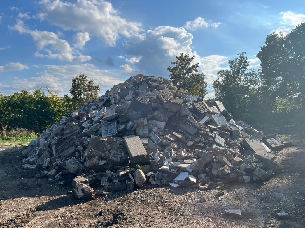 Ein großer Haufen aus Trümmern und Bruchsteinen liegt im Freien auf einer unbefestigten Fläche. Zwischen den Steinen sind zahlreiche Grabsteine und Fragmente mit eingravierten Inschriften erkennbar. Im Hintergrund stehen Bäume und Sträucher, darüber ein blauer Himmel mit vereinzelten Wolken.