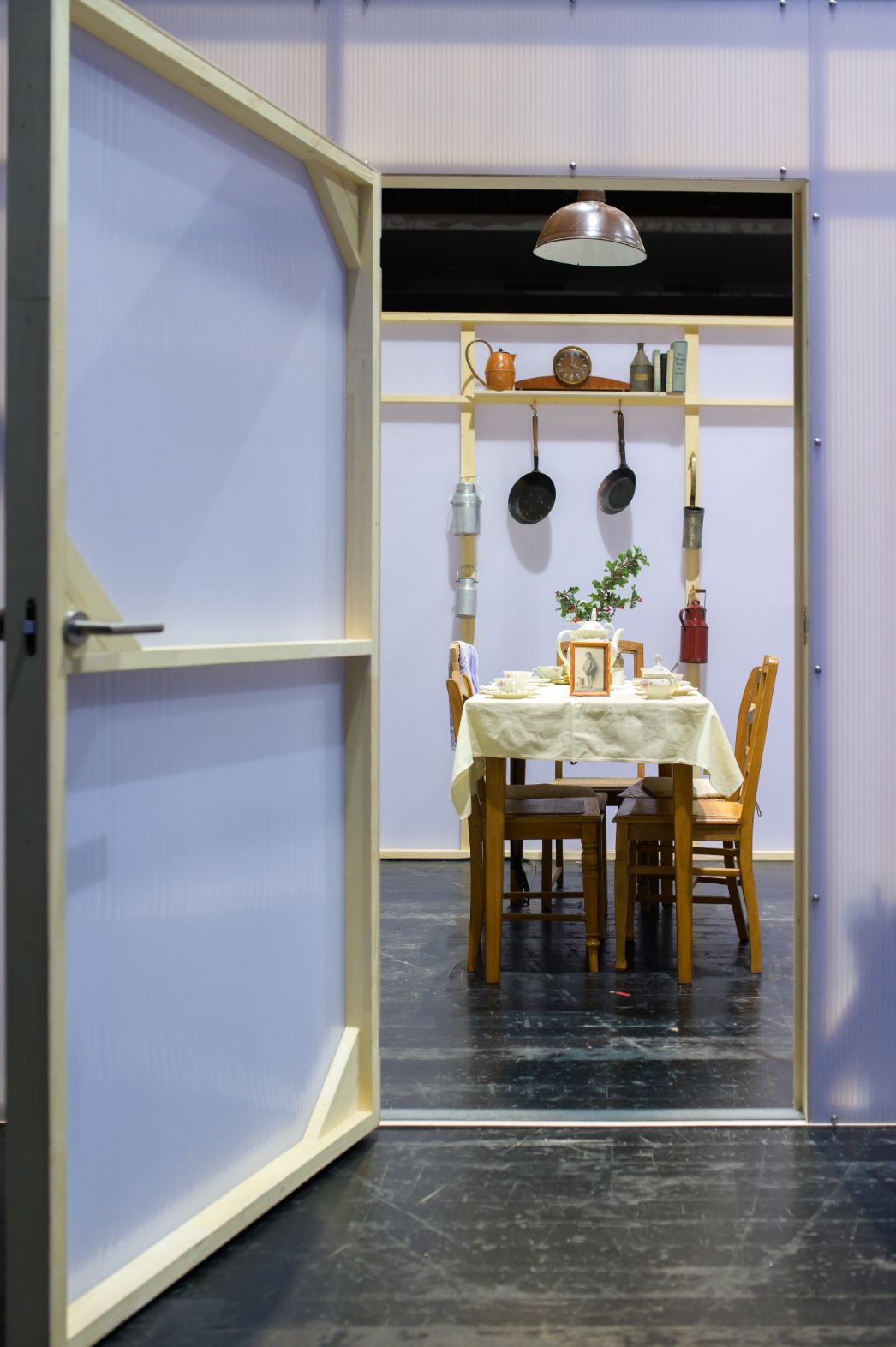 Ein begehbares Ausstellungsmodul inszeniert einen einfachen, gedeckten Küchentisch mit Alltagsgegenständen und einem persönlichen Foto. Die reduzierte, fast intime Raumgestaltung verweist auf häusliches Leben und individuelle Erinnerungen.