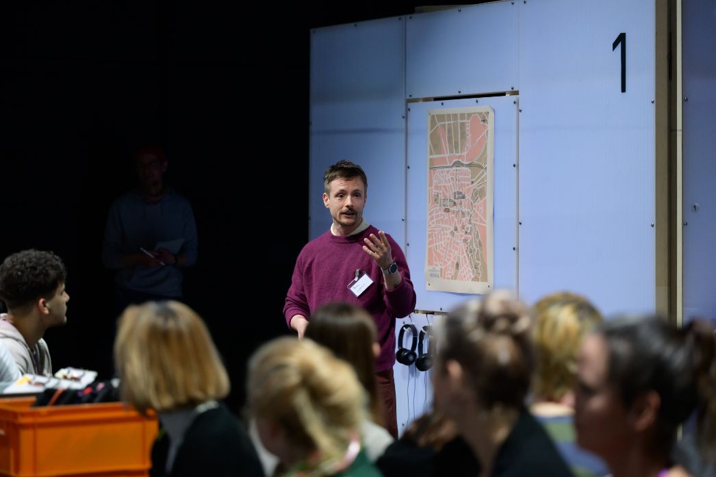 Ein Vermittler spricht vor einer Gruppe von Teilnehmenden in der Ausstellung und erläutert ein Exponat anhand einer Karte. Die Zuhörenden sitzen dicht beieinander und verfolgen aufmerksam die Ausführungen, während Kopfhörer auf eine mögliche vertiefende Audioebene hinweisen.