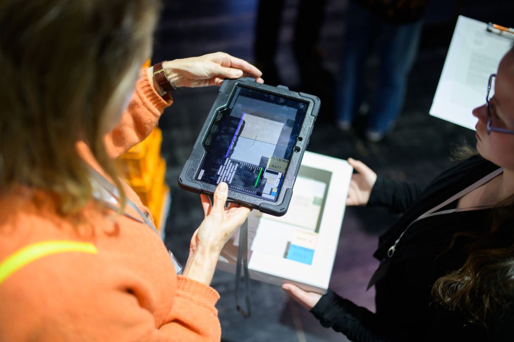 Eine Person zeigt auf einem Tablet digitale Inhalte zu einem Ausstellungsobjekt, während eine andere Teilnehmende begleitende Materialien in der Hand hält. Die Szene verdeutlicht das Zusammenspiel von analogen und digitalen Vermittlungsformaten.