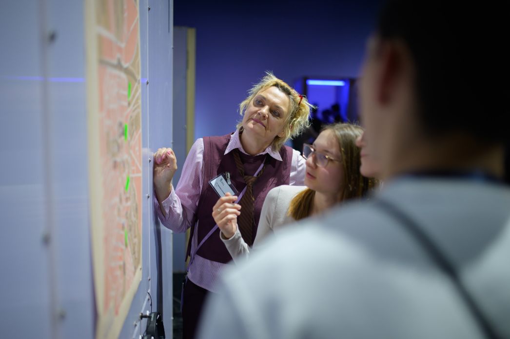 Eine Vermittlerin erläutert einer kleinen Gruppe ein Ausstellungsobjekt und verweist dabei auf Details einer Karte. Die Teilnehmenden verfolgen aufmerksam ihre Erklärungen und beteiligen sich am gemeinsamen Betrachten.