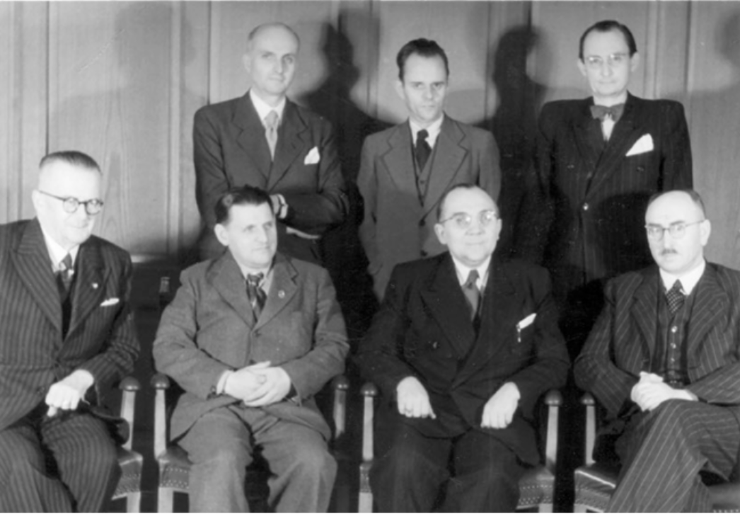 Schwarz-weiß-Gruppenporträt vom 5. Dezember 1946: Sieben ehemalige Buchenwald-Häftlinge in formeller Kleidung als Mitglieder der Thüringer Landesregierung. In der vorderen Reihe sitzen unter anderem Ministerpräsident Rudolf Paul (3. von links) sowie Innenminister und stellvertretender Ministerpräsident Ernst Busse (2. von links). Hinter ihnen stehen weitere Regierungsmitglieder, darunter Walter Wolf (2. von links), Volksbildungsminister. Die Aufnahme zeigt die frühe Nachkriegsregierung in offizieller Pose.