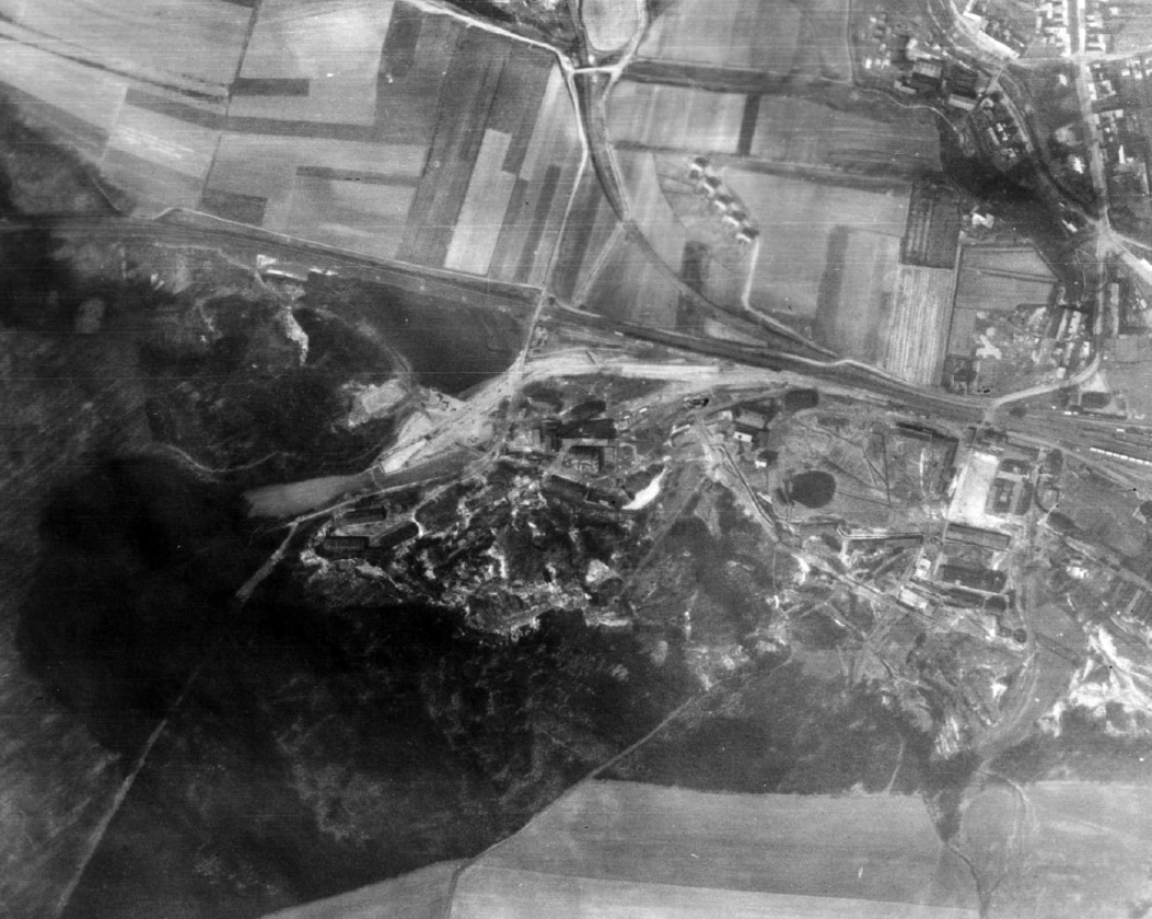 Schwarz-weiß-Luftaufnahme einer weitläufigen Landschaft mit Feldern, Straßen und einem größeren Lager- oder Industriekomplex. Mehrere Gebäude und Anlagen liegen am Rand eines Waldgebiets; Wege und Gleise verbinden das Gelände mit umliegenden Ortschaften.