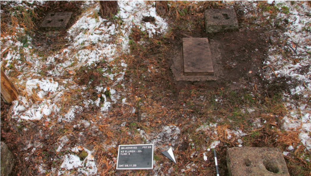 Fotografie eines Waldbodens mit archäologischen Befunden: Mehrere quadratische Betonfundamente mit Vertiefungen sind sichtbar, vermutlich Reste einer ehemaligen Lagerstruktur. Zwischen Laub, Erde und Schneeresten liegt eine beschriftete Tafel mit Angaben zur Fundstelle („KZ Ellrich-Juliushütte, Turm 3“) sowie ein kleiner Markierungspfeil.