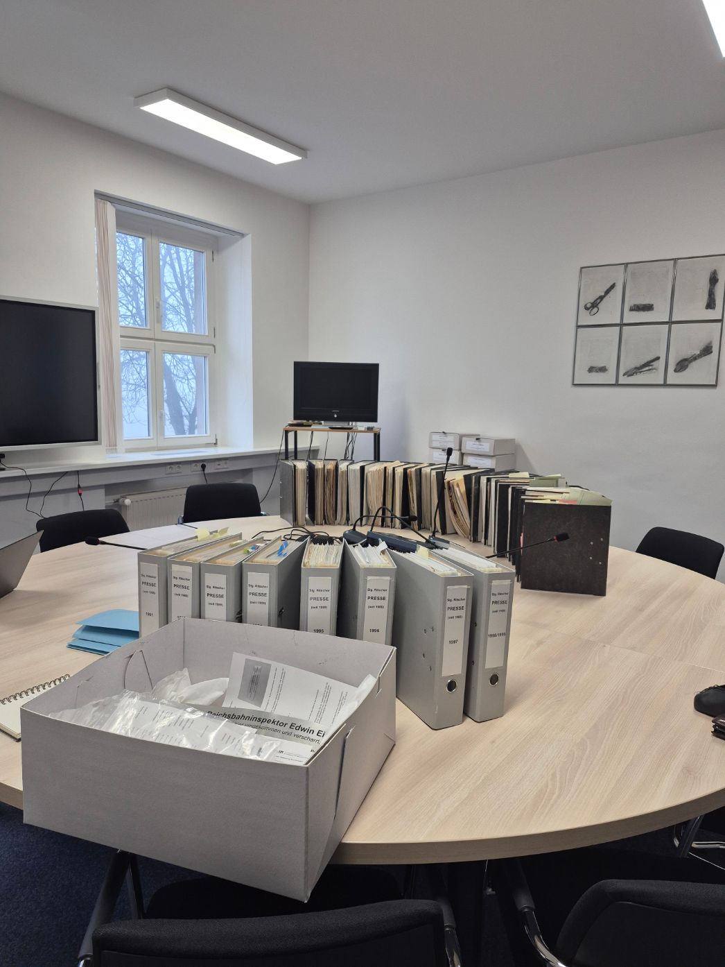 Innenraum eines hellen Arbeits- oder Archivraums mit einem großen runden Tisch. Auf dem Tisch stehen zahlreiche Ordner, Aktenmappen und Dokumentenkästen, teilweise beschriftet und nach Jahren sortiert. Mikrofone sind am Tisch installiert, was auf Besprechungen oder Forschungsarbeit hinweist. Im Hintergrund befinden sich Fenster, zwei Monitore und weitere Aktenbestände. An der Wand hängen gerahmte Abbildungen.