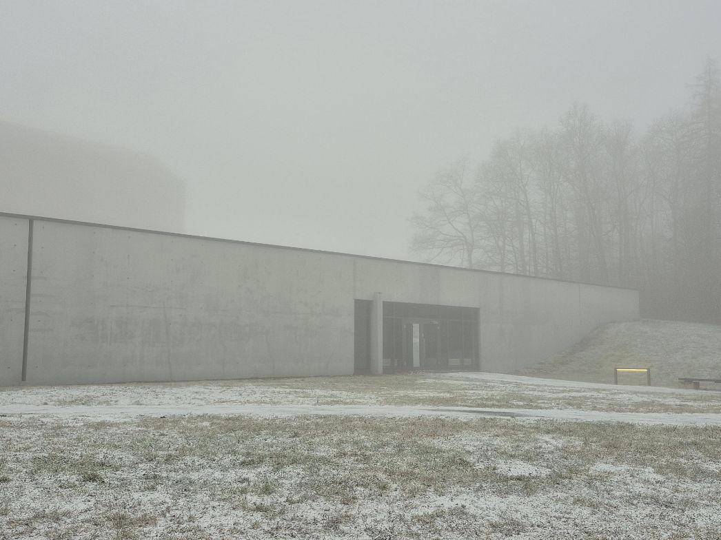 Flaches, modernes Ausstellungsgebäude aus Beton mit klaren Linien, teilweise in den Hang gebaut. Der Eingang liegt mittig und ist durch eine Glasfront markiert. Die Umgebung ist von dichtem Nebel verhüllt, wodurch Bäume im Hintergrund nur schemenhaft zu erkennen sind. Der Boden ist leicht mit Schnee bedeckt, und die gesamte Szene wirkt ruhig und gedämpft.