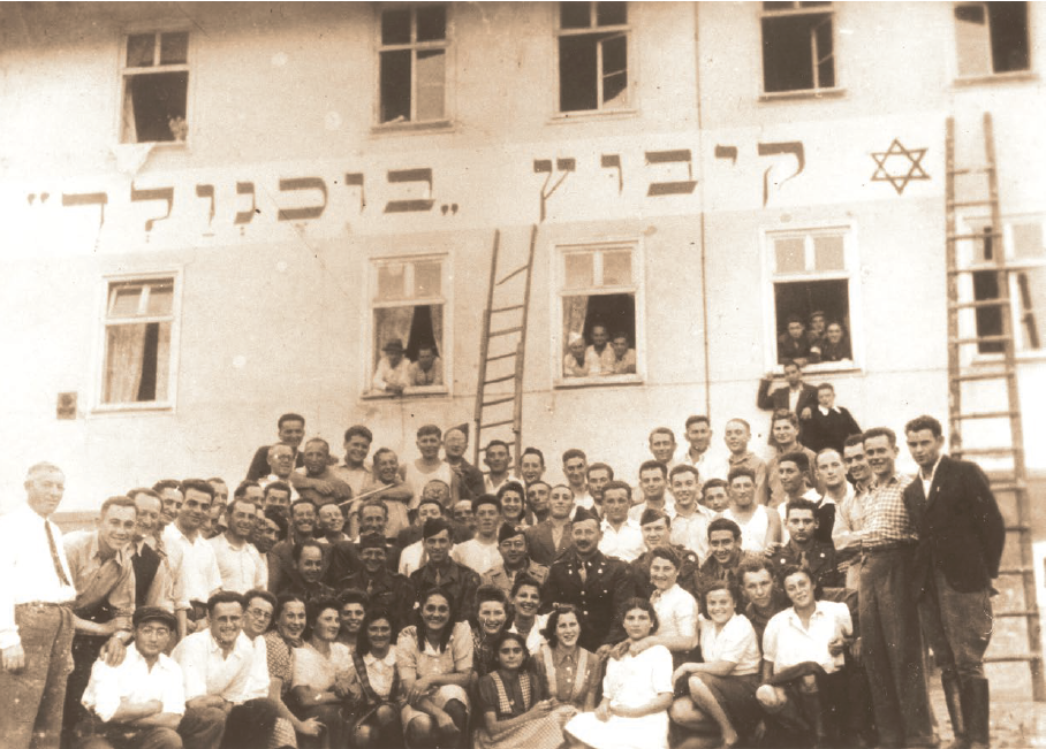 Historisches Gruppenfoto vor einem Gebäude: Zahlreiche junge Menschen stehen und sitzen dicht gedrängt zusammen und blicken in die Kamera. An der Fassade über ihnen befindet sich eine hebräische Inschrift sowie ein Davidstern; einige Personen schauen aus geöffneten Fenstern, und Leitern lehnen an der Hauswand.