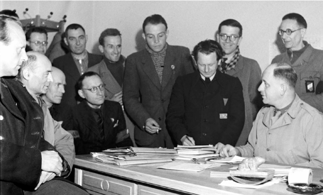 Schwarz-weiß-Fotografie vom 24. April 1945: Mitglieder des Internationalen Lagerkomitees versammeln sich im ehemaligen Arbeitszimmer des Lagerkommandanten im KZ Buchenwald. Am Schreibtisch sitzt der Übergangskommandant der US-Armee, Lorenz Schmuhl. Links neben ihm der deutsche politische Häftling Walter Bartel. Im Hintergrund, ganz links stehend mit Brille, der deutsche politische Häftling Harry Kuhn. Auf dem Tisch liegen Aktenstapel; die Szene zeigt eine administrative Besprechung nach der Befreiung.