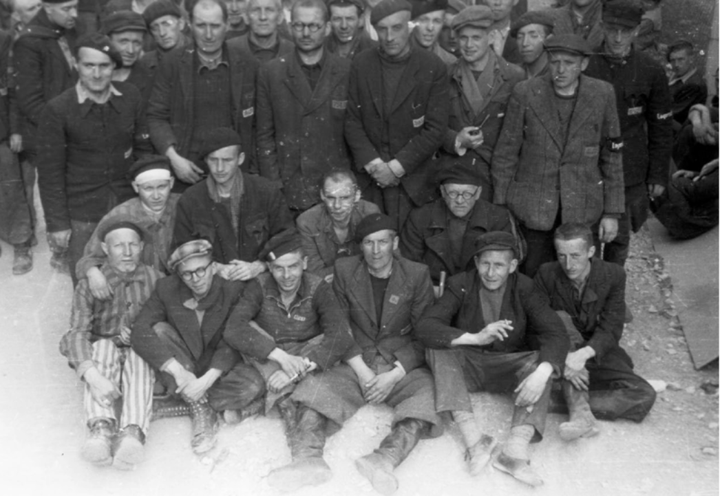 Schwarzweiß-Gruppenfotografie von etwa 25 männlichen Häftlingen in einem Konzentrationslager. Die Männer tragen einfache, teils zerlumpte Kleidung, einige Häftlingsjacken und Mützen; einer in der vorderen Reihe trägt gestreifte Lagerkleidung. Viele wirken abgemagert und erschöpft. Sie sind dicht zusammengedrängt, einige sitzen am Boden, andere stehen dahinter. Mehrere blicken direkt in die Kamera, ihre Gesichtsausdrücke sind ernst bis müde. Die Aufnahme vermittelt die Enge und die Belastung des Lageralltags.