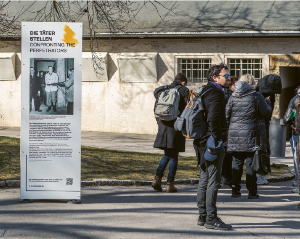 Eine freistehende Stele mit der Überschrift „Die Täter / Confronting the Perpetrators“ steht im Außenbereich vor einem Gebäude. Darauf sind ein historisches Foto und erklärender Text zu sehen. In der Umgebung bewegen sich mehrere Besucher:innen über das Gelände der Gedenkstätte.