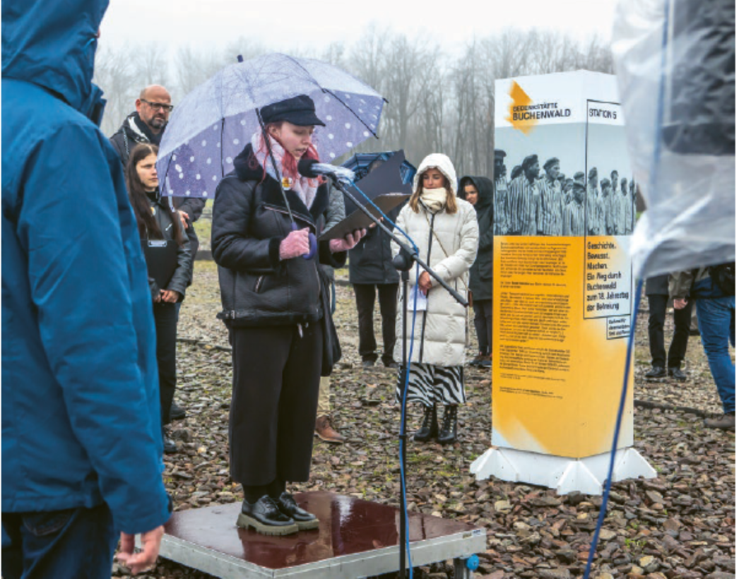 Eine Frau steht bei Regen unter einem Regenschirm auf einer kleinen Plattform und liest aus einem Tablet in ein Mikrofon. Um sie herum stehen mehrere Zuhörer:innen. Neben ihr befindet sich eine freistehende Stele mit Texten und historischen Bildern zur Gedenkstätte Buchenwald.