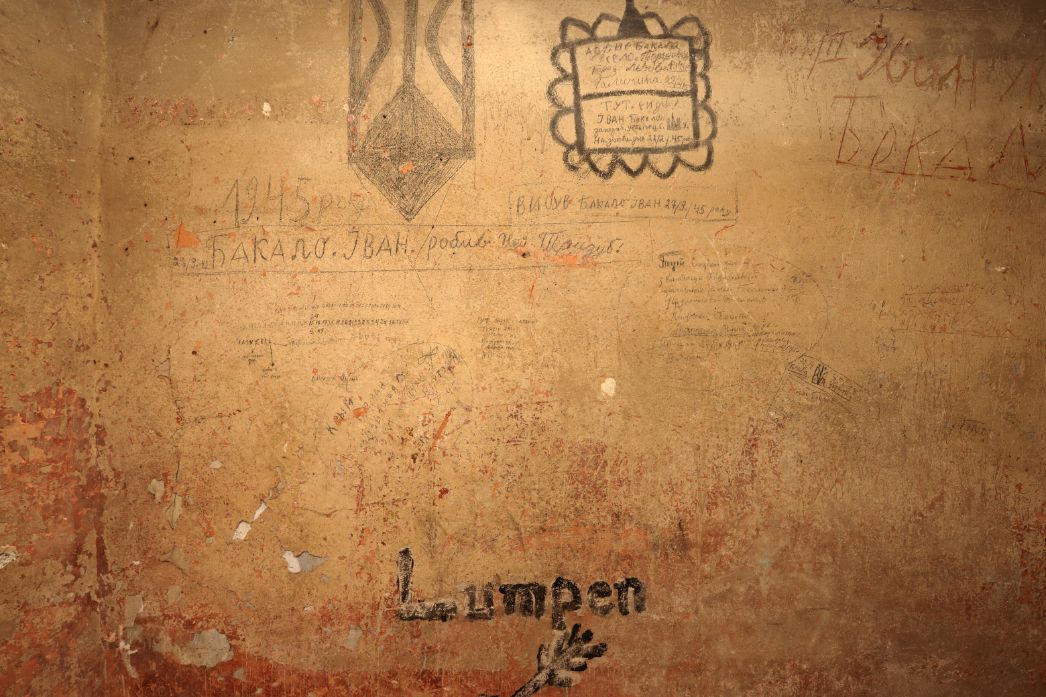 Stark verwitterte Innenwand mit bröckelndem Putz in beige- und rötlichen Farbtönen. Über die Fläche verteilt befinden sich zahlreiche eingeritzte und gezeichnete Inschriften, vor allem in kyrillischer Schrift, darunter Namen, Daten und kurze Botschaften. Oben sind mehrere Zeichnungen zu sehen, darunter ein trident-ähnliches Symbol und ein eingerahmter Text. In der unteren Bildhälfte steht in dunkler Schrift deutlich das Wort „Lumpen“, darunter eine kleine Zeichnung, möglicherweise eine Hand oder ein Zweig mit Blättern.