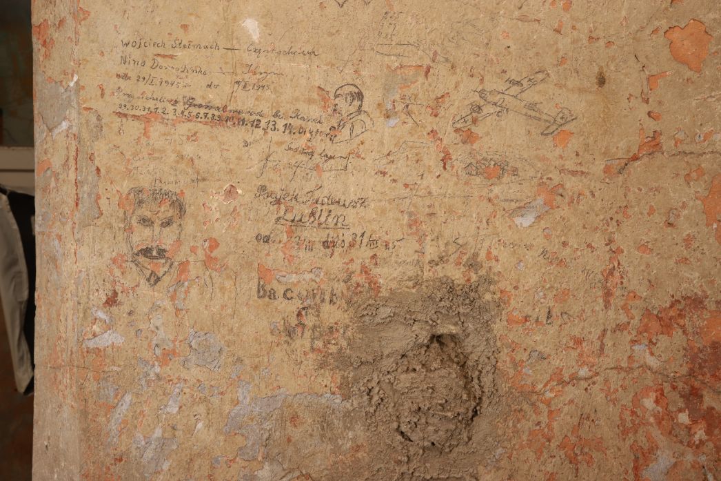 Detail einer stark verwitterten Innenwand mit abgeplatztem Putz in Beige- und Rottönen. Auf der Oberfläche sind zahlreiche eingeritzte oder mit Bleistift geschriebene Notizen, Namen und Daten zu erkennen, darunter auch kleine Zeichnungen wie Gesichter und ein Flugzeug. Unten mittig befindet sich ein größerer, unregelmäßiger Fleck aus rauem, aufgetragenem Material. Links am Bildrand ist ein Teil eines Türdurchgangs mit hängendem Stoff zu sehen.