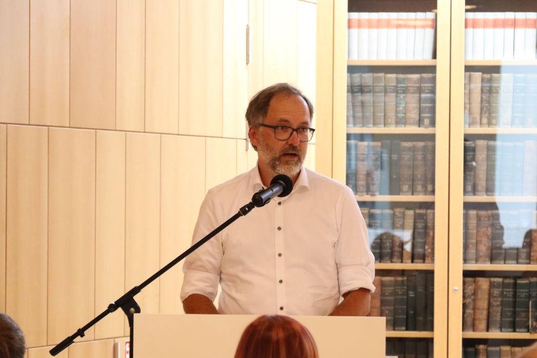 Ein Mann mittleren Alters mit Brille und grauem Bart steht an einem Rednerpult und spricht in ein Mikrofon. Er trägt ein weißes Hemd und blickt leicht zur Seite, als würde er sich an ein Publikum wenden. Im Hintergrund sind helle Holzschränke sowie ein verglastes Bücherregal mit zahlreichen alten, gebundenen Büchern zu sehen. Im Vordergrund ist unscharf der Kopf einer Zuhörerin oder eines Zuhörers erkennbar.