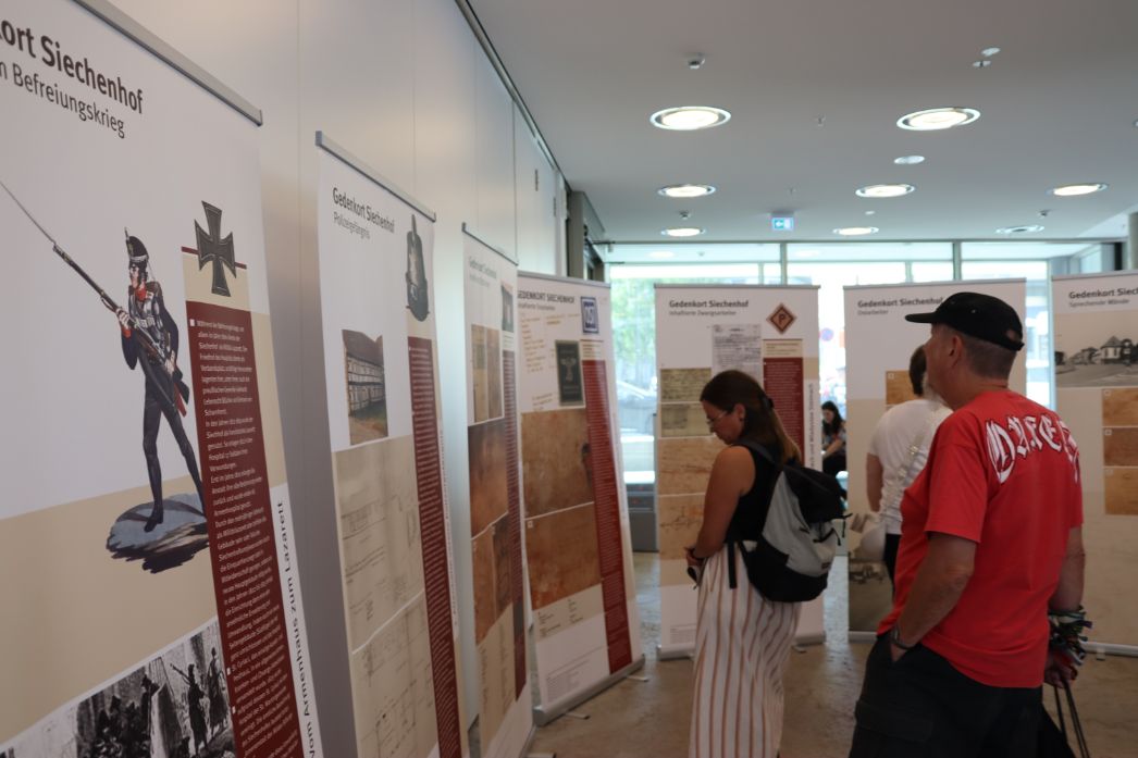 Innenraum einer Ausstellung mit mehreren freistehenden Informationstafeln. Die Tafeln zeigen Texte, historische Abbildungen und Dokumente zum Thema „Gedenkort Siechenhof“. Zwei Besucher stehen davor und lesen: eine Person mit Rucksack und eine weitere Person im roten T-Shirt und schwarzer Kappe. Der Raum ist hell, mit großen Fenstern im Hintergrund und runden Deckenleuchten.