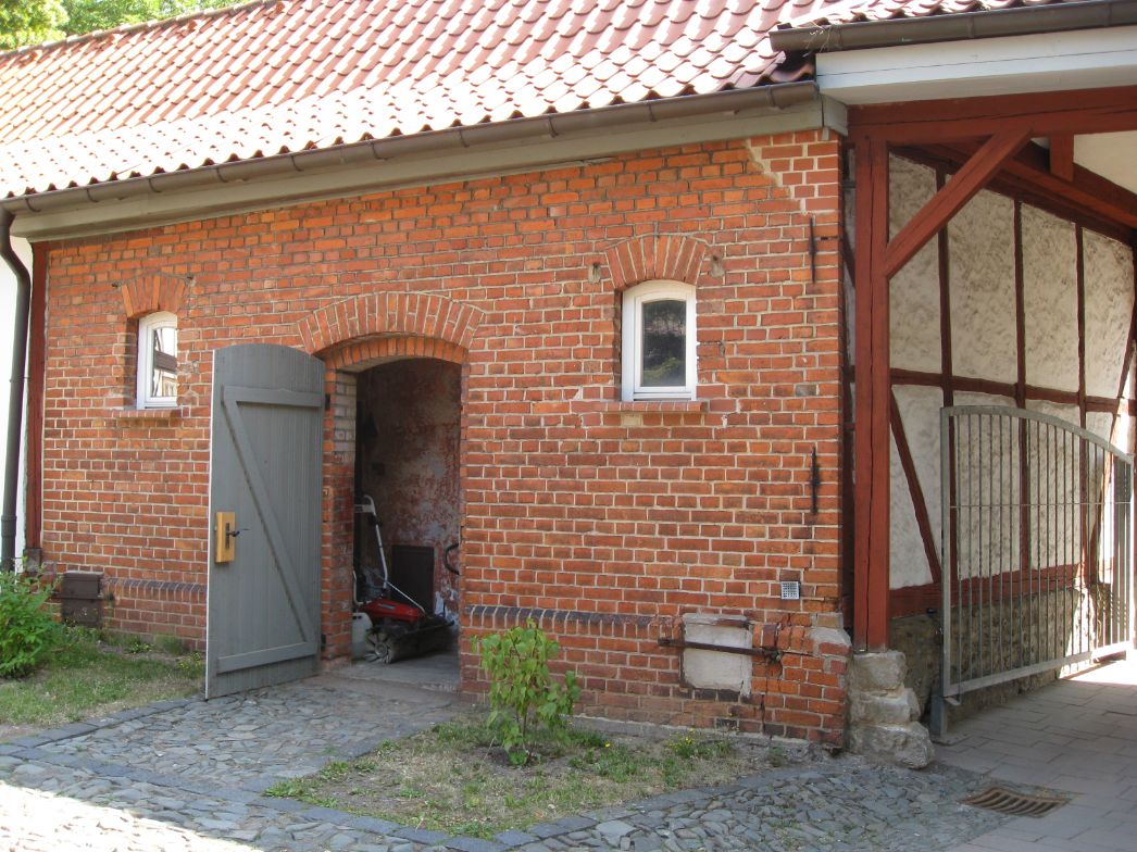 Backstein-Nebengebäude mit rotem Ziegeldach. In der Mitte befindet sich ein offenes, rundbogiges Tor mit geöffneter grauer Holztür; im Inneren ist ein Rasenmäher zu sehen. Links und rechts davon kleine, schmale Fenster mit weißen Rahmen. Rechts schließt ein Fachwerk-Anbau mit Holzbalken und weiß verputzten Flächen sowie einem Metalltor an. Vor dem Gebäude liegen Kopfsteinpflaster und etwas spärliche Vegetation.