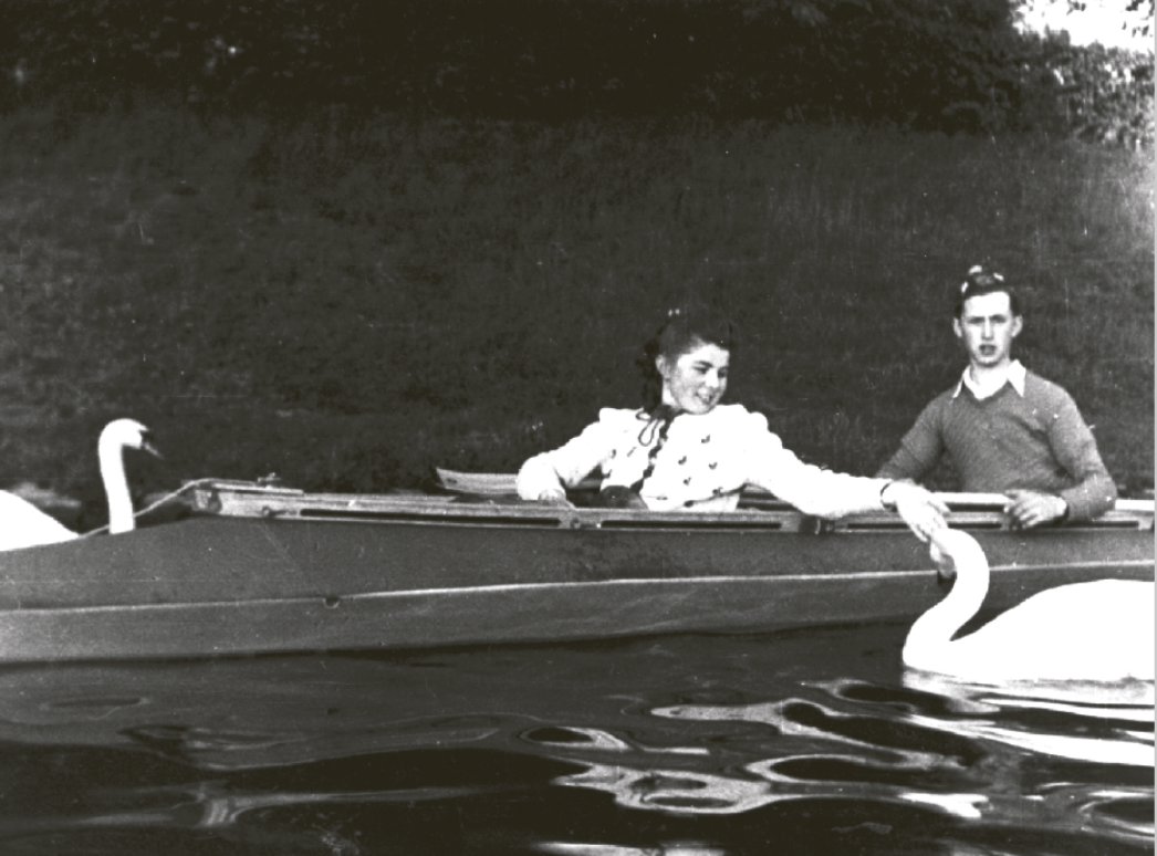 Historisches Schwarzweißfoto von zwei Jugendlichen in einem Ruderboot auf einem See. Eine junge Frau beugt sich nach vorne und füttert einen Schwan, während ein junger Mann neben ihr sitzt und in die Kamera schaut.