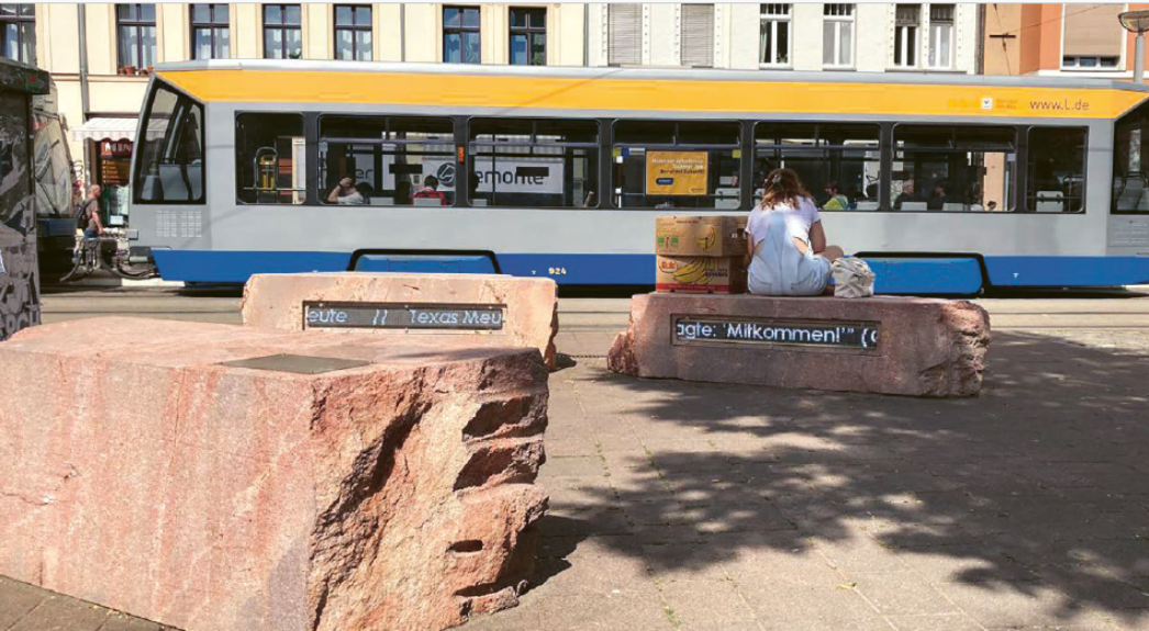 Drei große Steinblöcke im öffentlichen Raum, die als Gedenkzeichen gestaltet sind, stehen auf einem Platz. Auf ihnen befinden sich eingelassene Textbänder. Im Hintergrund fährt eine Straßenbahn vorbei, und eine Person sitzt auf einem der Steine.