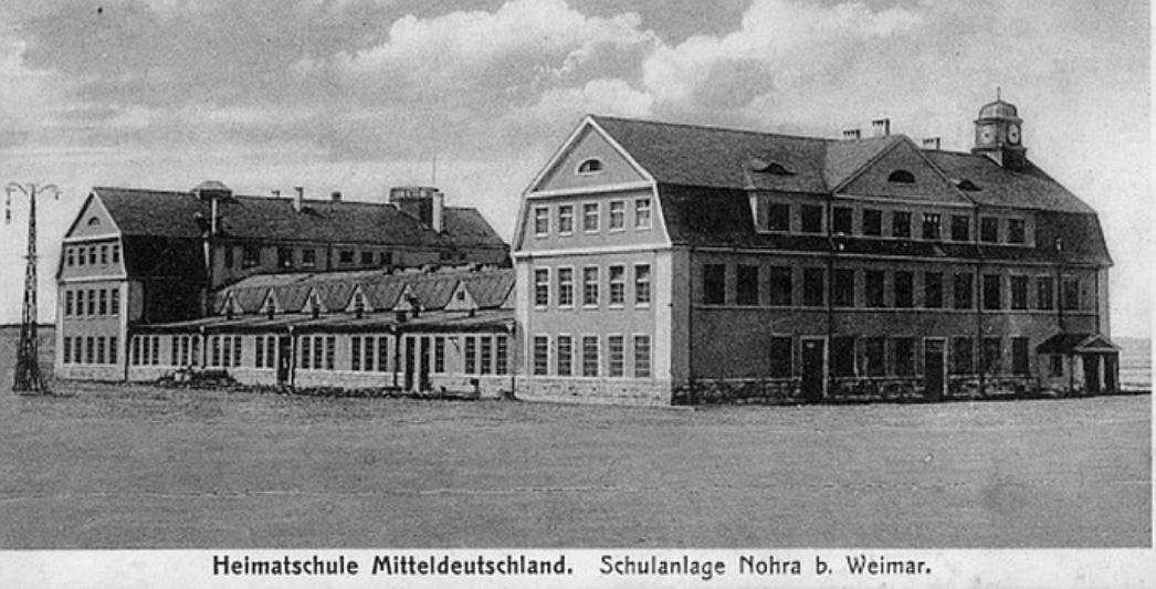  Ausschnitt aus einer historischen Postkarte. Sie zeigt ein großes Gebäude mit zwei höheren Flügeln, die in der Mitte durch einen flachen Teil verbunden sind. Darunter steht "Heimatschule Mitteldeutschland. Schulanlage Nohra bei Weimar"