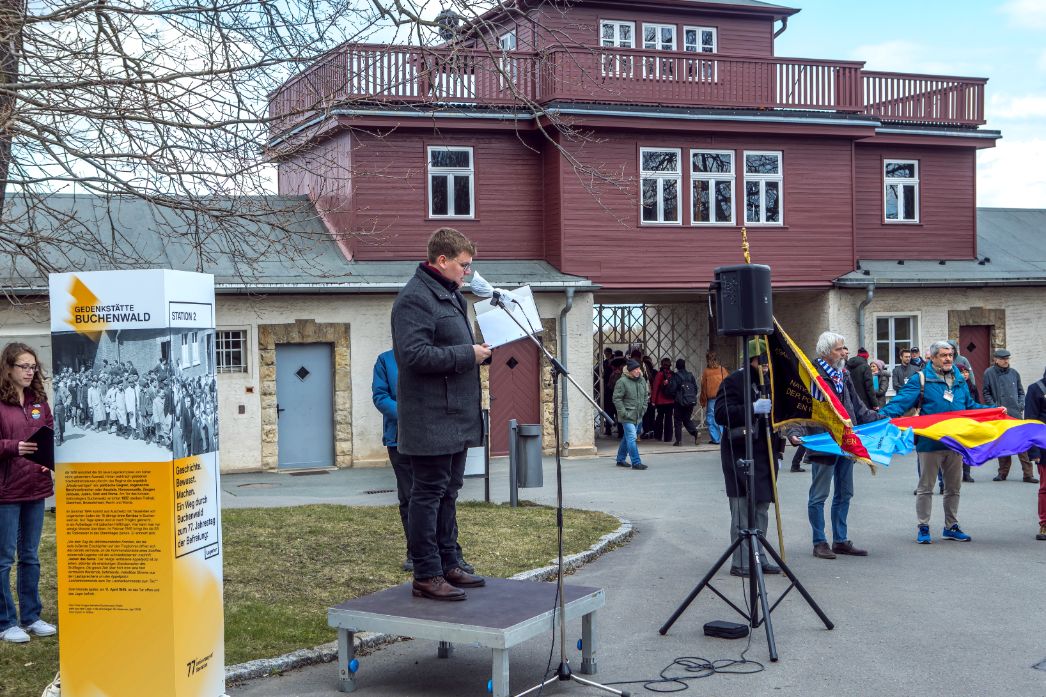 In the center of the picture, a speaker with a black jacket stands on a small stage element and speaks into a microphone. To his left is a gray-white-orange information stele. In the background, the gate building of the former concentration camp can be seen.