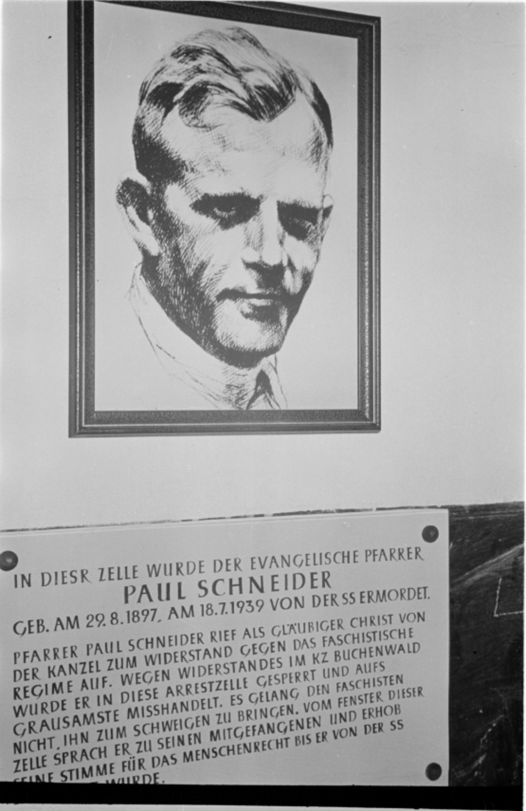 Zu sehen ist ein Portrait eines Mannes an einer Wand. Darunter wurde eine Gedenkstafel angebracht. Auf der tafel steht: "In dieser Zelle wurde der evangelische Pfarrer Paul Schneider Geb, am 29.8.1897 am 18.7.1939 von der SS ermordet.