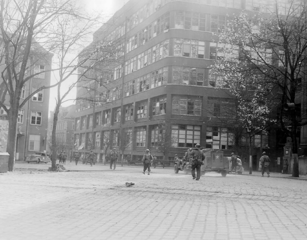 Schwarz-weiß-Fotografie einer deutschen Stadt im Frühjahr 1945: US-amerikanische Soldaten bewegen sich mit einem Militärfahrzeug durch eine Straßenkreuzung vor einem mehrstöckigen Gebäude. Die Szene zeigt die Besetzung einer Stadt unmittelbar nach dem Einmarsch der alliierten Truppen.