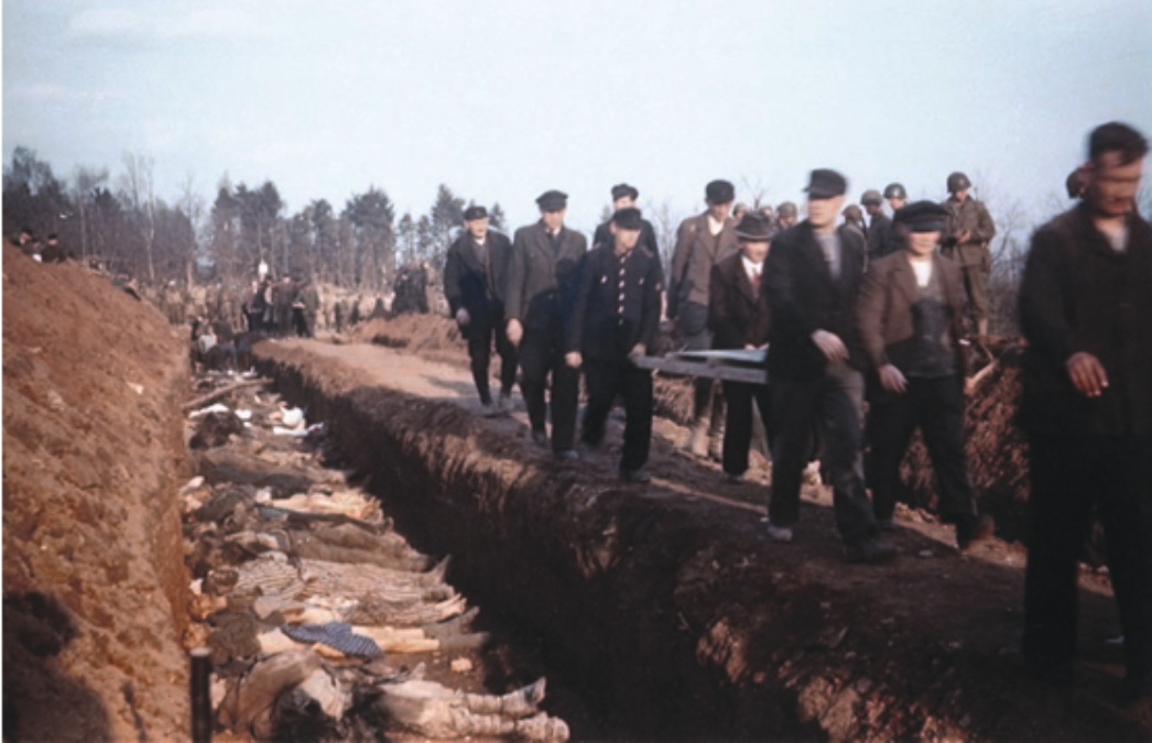 Eine Gruppe von Männern geht entlang eines langen Massengrabens, in dem zahlreiche Leichen nebeneinander liegen. Die Szene wirkt dokumentarisch und zeigt offenbar Zivilisten, die unter Aufsicht das Ausmaß von NS-Verbrechen unmittelbar nach der Befreiung eines Konzentrationslagers besichtigen.