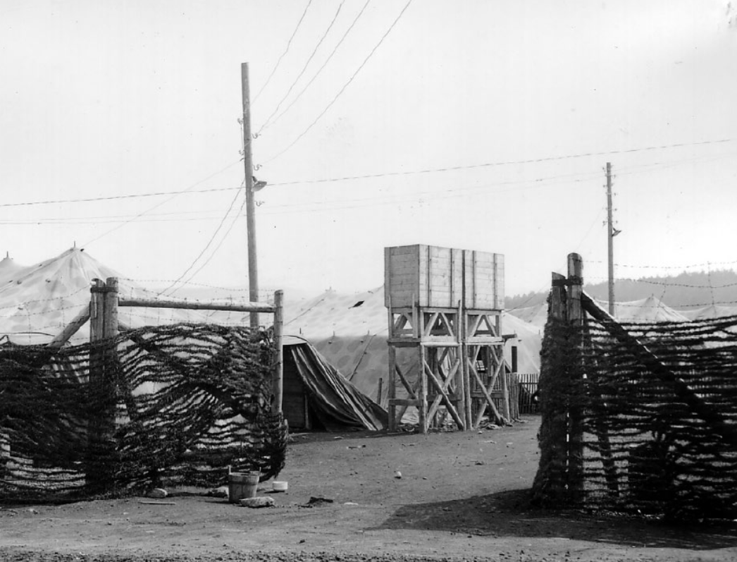 Schwarz-weiß-Fotografie eines eingezäunten Lagerbereichs: Ein Eingang aus Holzpfosten und Stacheldraht führt zu mehreren Zelten. Im Hintergrund steht ein hölzerner Wasserturm auf Stelzen, daneben verlaufen Stromleitungen. Die Anlage wirkt provisorisch und erinnert an ein Lager für Gefangene oder Zwangsarbeiter.