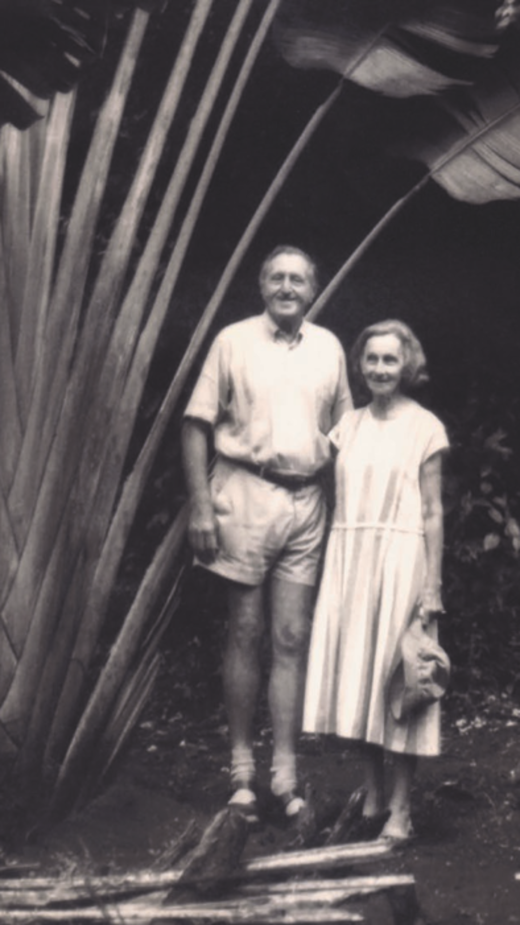 Schwarzweißfotografie eines älteren Paares, das nebeneinander im Freien steht. Der Mann trägt ein kurzärmeliges Hemd und kurze Hose, die Frau ein helles, knielanges Kleid und hält einen Gegenstand in der Hand. Beide lächeln leicht in die Kamera. Im Hintergrund und seitlich rahmen große tropische Pflanzen mit breiten Blättern die Szene ein, was auf eine Aufnahme in einer warmen, möglicherweise kolonialen Umgebung hindeutet.