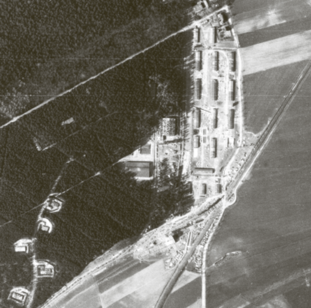 Schwarz-weiß-Luftaufnahme eines Industrie- und Lagergeländes am Waldrand: Mehrere langgestreckte Gebäude liegen parallel zueinander, umgeben von Straßen, Gleisen und freien Flächen. Die Anlage wirkt abgeschieden und ist von Feldern sowie dichtem Wald umgeben.