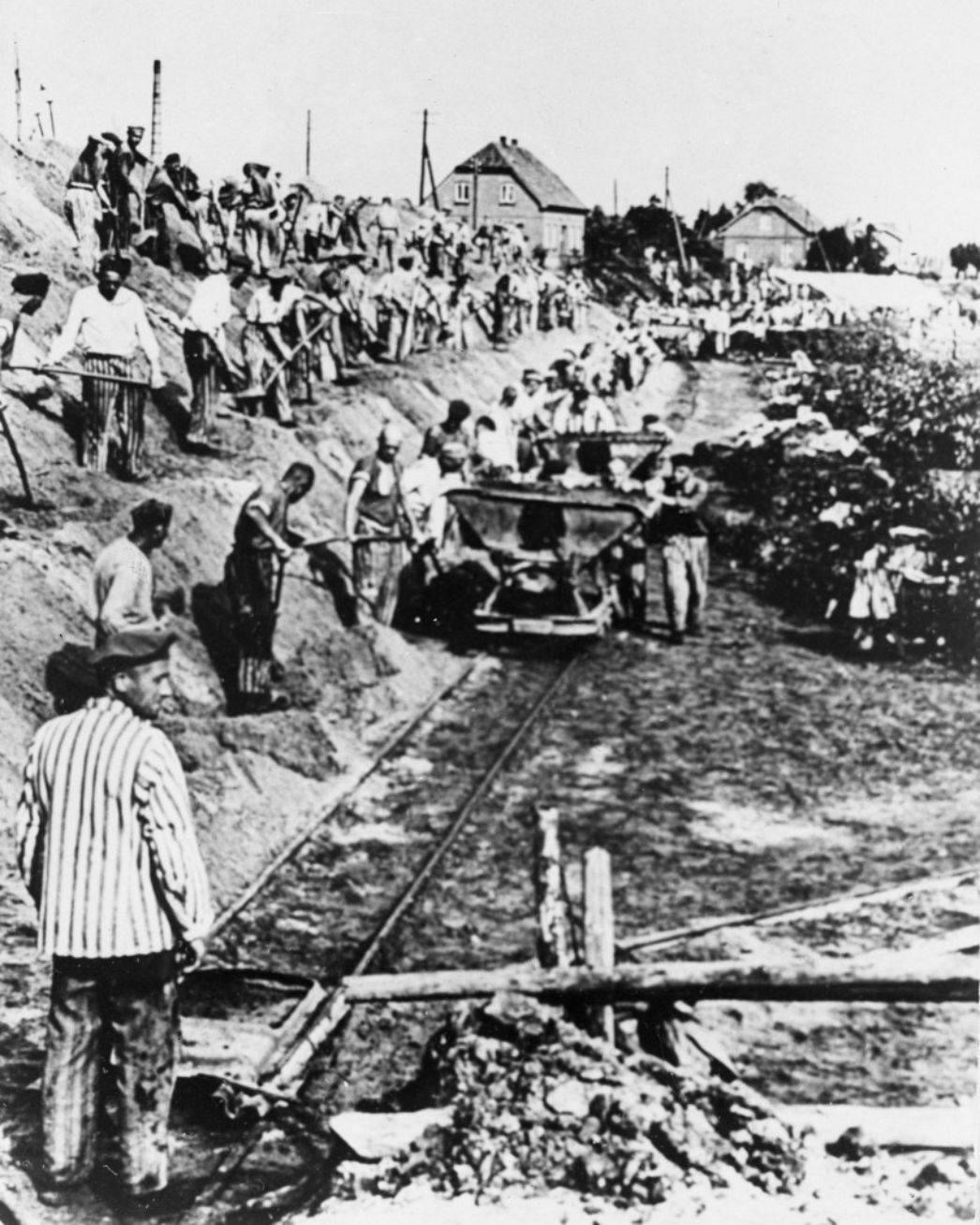 Das Bild zeigt Zwangsarbeiter die entlang einer Lorentrasse mit Schaufelarbeiten beschäftigt sind. Auf halber Strecke der Trasse steht eine Kipplore auf den Gleisen.