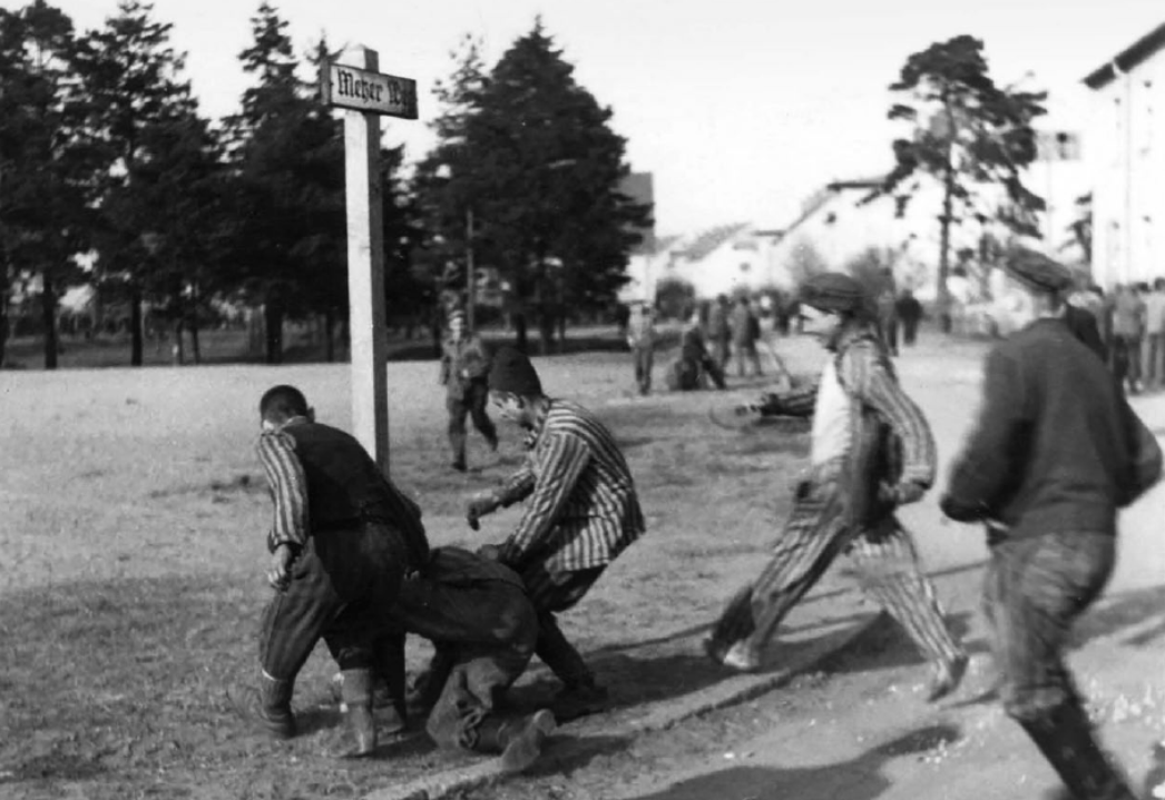 Historisches Schwarzweißfoto: Mehrere Häftlinge in gestreifter Kleidung schlagen oder treten eine am Boden liegende Person. Im Hintergrund sind weitere Menschen sowie Gebäude und Bäume zu sehen.
