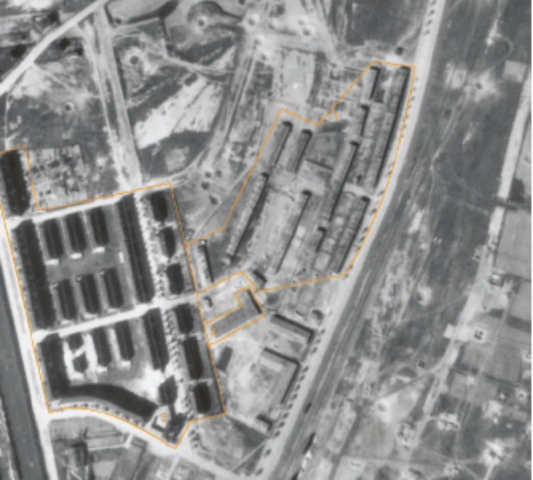 Schwarz-weiß-Luftaufnahme eines größeren Lager- und Industriekomplexes. Mehrere Baracken und Gebäude sind in Blöcken angeordnet und durch eine orangefarbene Linie hervorgehoben. Das Gelände ist von Straßen, Gleisen und freien Flächen umgeben und zeigt die Struktur eines Konzentrationslager-Außenlagers oder eines zugehörigen Produktionsstandorts.