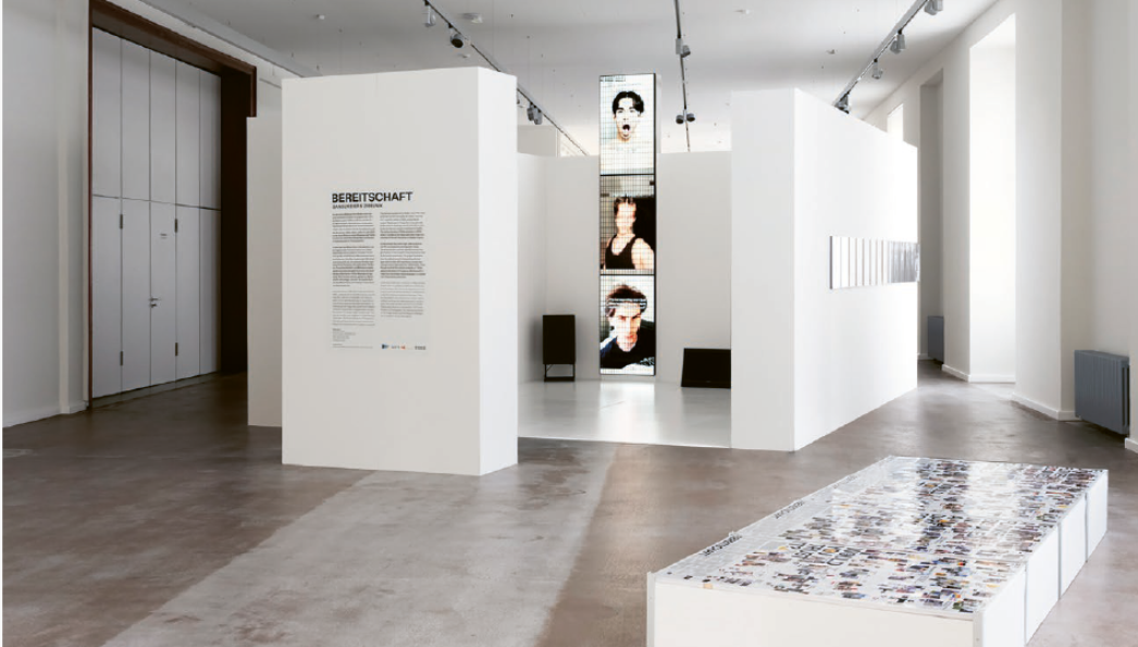 Ausstellungsraum eines Museums mit weißen Stellwänden und klarer, moderner Gestaltung. Auf einer Wand ist der Titel „Bereitschaft“ mit Begleittext zu sehen. In der Mitte steht eine schmale, hohe Videoinstallation mit mehreren Bildsequenzen von Gesichtern. Im Vordergrund befindet sich eine niedrige Vitrine oder Plattform mit vielen ausgelegten Bildern oder Dokumenten.